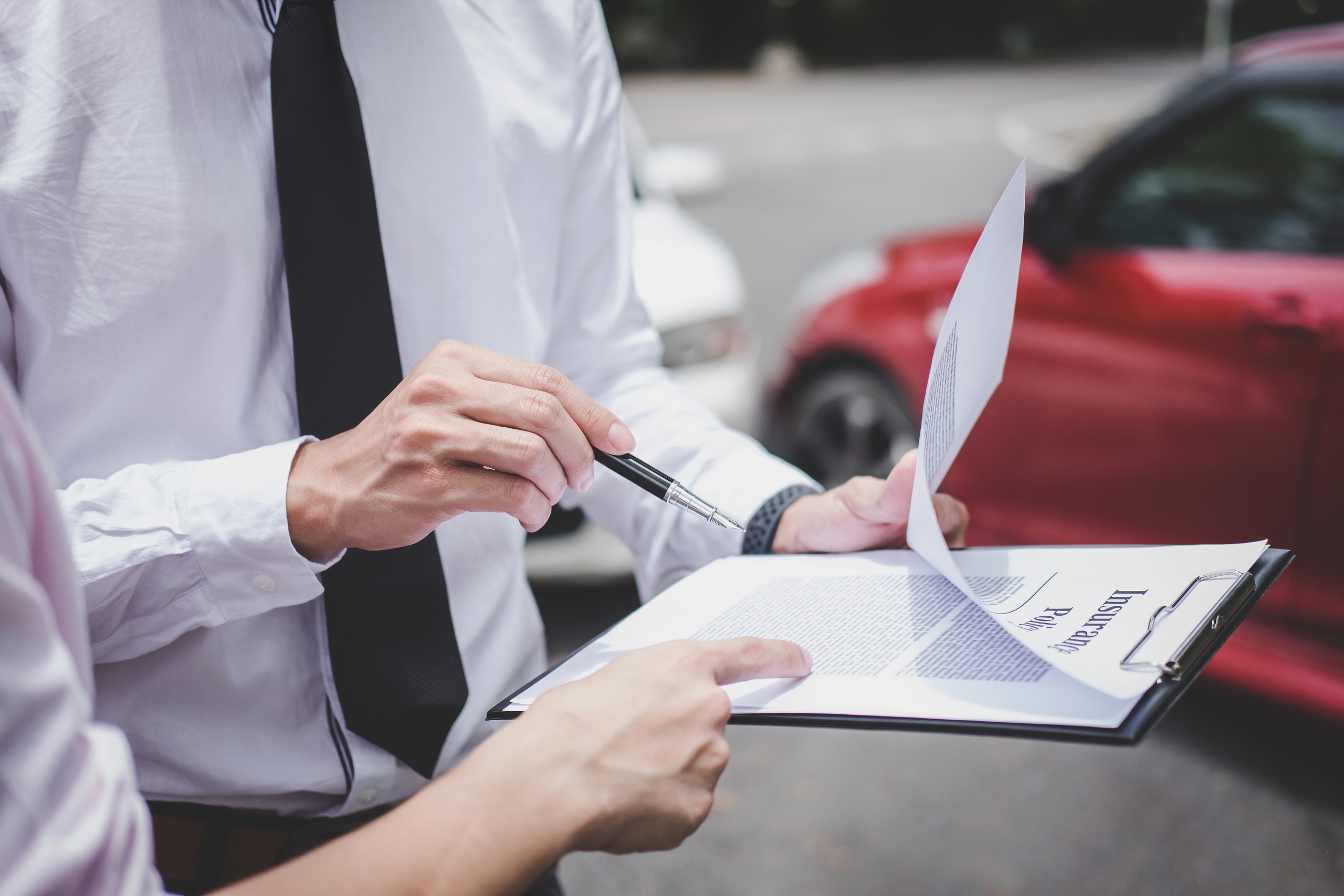 Two people review an insurance policy document near parked cars.