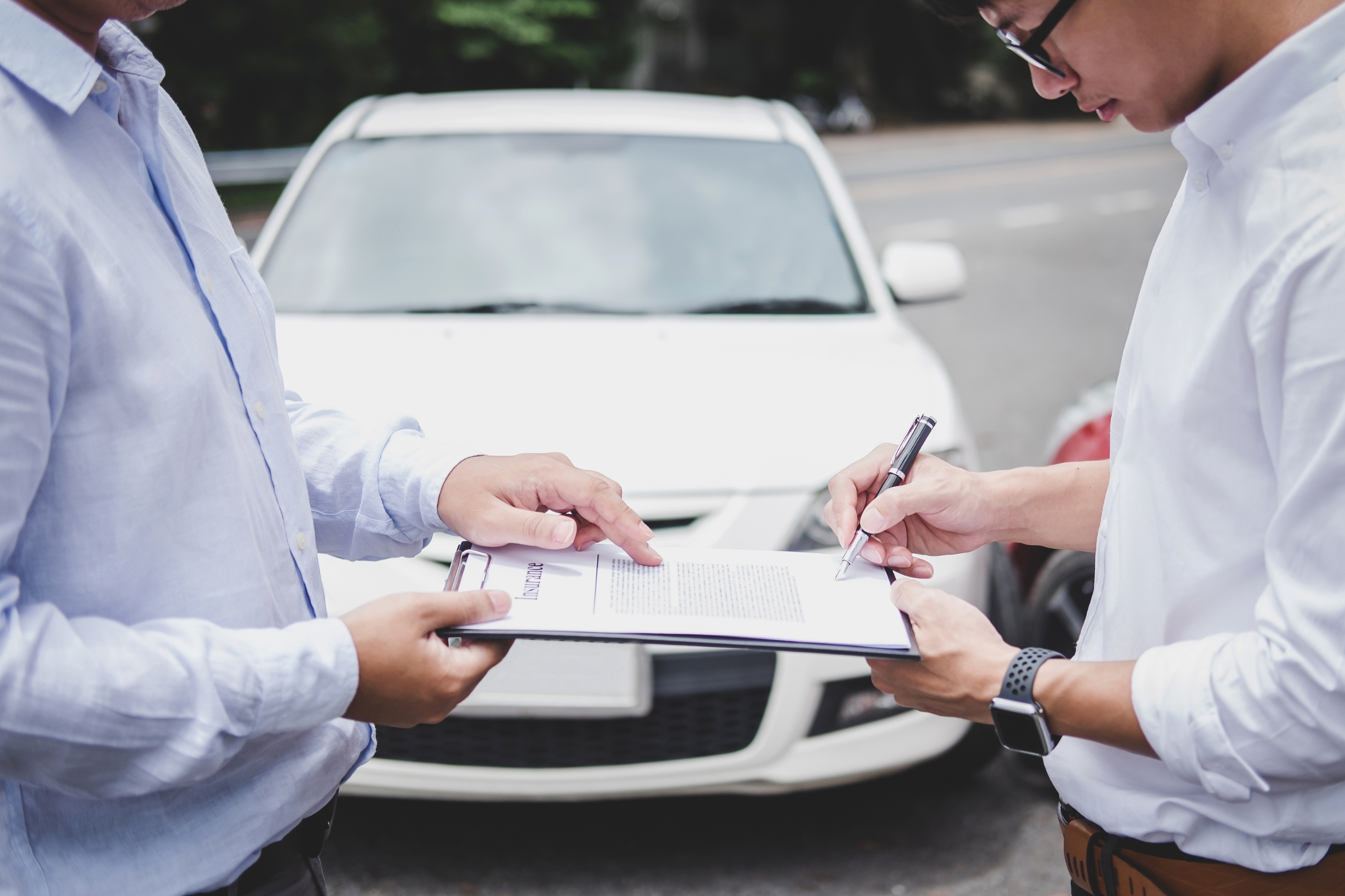 Two men signing an insurance document in front of a white car.