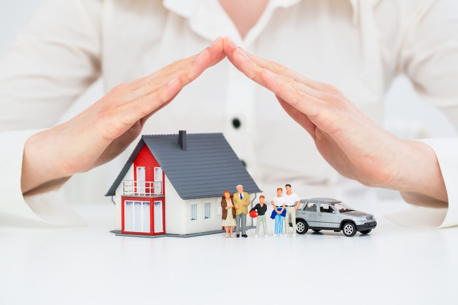 Hands protect a miniature house, family figures, and a car, symbolizing security.